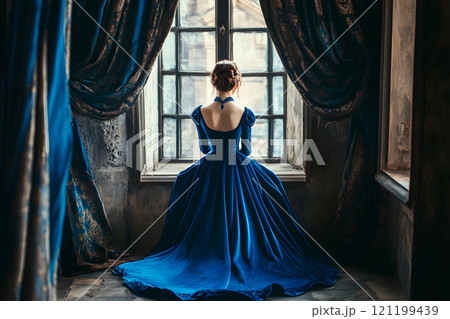 Woman in sapphire-blue gown gazes out window of a vintage interior. 121199439