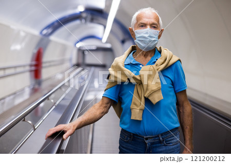 Elderly man in protective mask moving up on escalator while leaving subway station platform 121200212