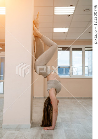 The girl is training in the yoga studio. Dressed in a beige tracksuit doing a handstand against the wall. Healthy lifestyle and yoga concept. The girl is training in the yoga studio. Dressed in a beige tracksuit doing a handstand against the wall. Healthy lifestyle and yoga concept. 121200746