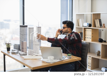 Indian male architect in his 30s discussing city model design during phone call. Focused and professional atmosphere with architectural elements and modern office setting. Indian male architect in his 30s discussing city model design during phone call. Focused and professional atmosphere with architectural elements and modern office setting. 121201397
