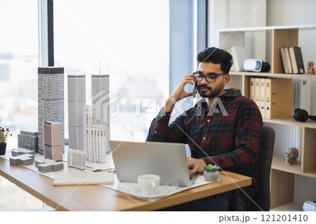 Young Indian male architect sitting at desk with laptop and scale models, talking on phone, focused on designing modern city buildings. Young Indian male architect sitting at desk with laptop and scale models, talking on phone, focused on designing modern city buildings. 121201410