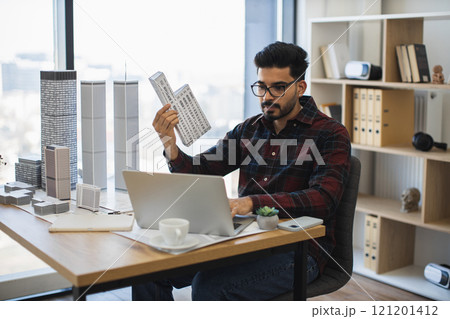 Indian male architect in twenties working on city model at office desk with modern buildings and laptop. 121201412