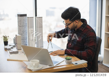 Indian male architect in modern office measures city model showcasing creativity and precision in design process, highlighting architectural skills. 121201439