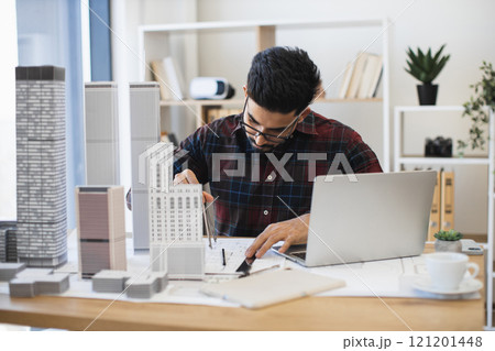 Indian male architect drawing modern city plan with pencil and paper. Young professional working on architectural project with scale models and laptop in studio. 121201448