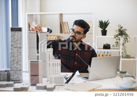 Indian male architect in his 30s creating detailed plan for modern urban city using building models. Workspace has laptop and architectural materials indicating creativity and innovation. 121201456