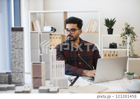 Young Indian male architect working on laptop designing modern urban development using building models. Office setting with books and plants in background, reflecting focus and creativity. Young Indian male architect working on laptop designing modern urban development using building models. Office setting with books and plants in background, reflecting focus and creativity. 121201458