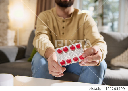 Young sick, feels badly man in casual beige shirt holds blister pack of red lozenges, sitting in bright, cozy living room. Young sick, feels badly man in casual beige shirt holds blister pack of red lozenges, sitting in bright, cozy living room. 121203409