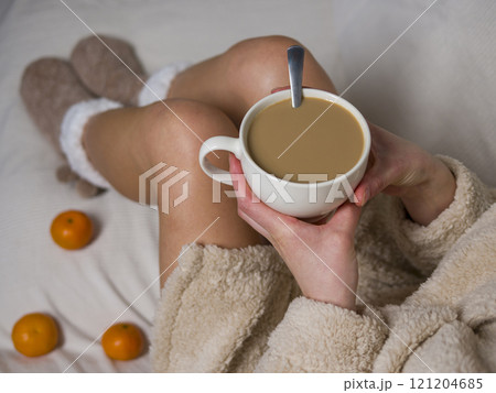 Woman in beige bathrobe and fluffy slippers enjoying a cozy morning at home with a cup of coffee and tangerines in bed Woman in beige bathrobe and fluffy slippers enjoying a cozy morning at home with a cup of coffee and tangerines in bed 121204685