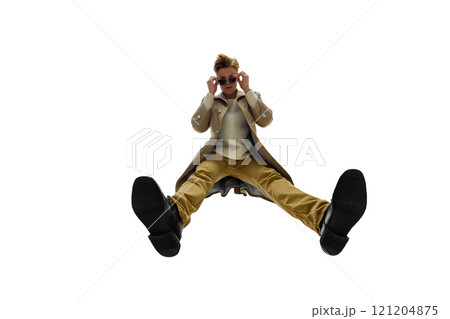 Modern young man in beige trench coat and black boots captured in striking pose against white background. Bottom view. Stylish appearance. 121204875