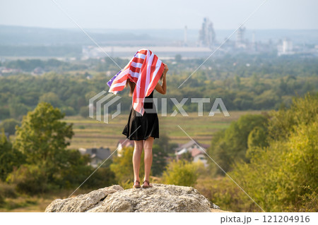 Young happy woman with long hair raising up waving on wind american national flag in her hands standing on high rocky hill enjoying warm summer day. 121204916