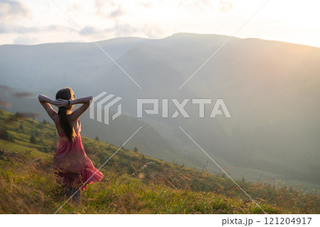 Young happy woman traveler in red dress standing on grassy hillside on a windy evening in summer mountains enjoying view of nature at sunset. 121204917