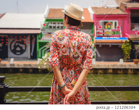 Young woman in ethnic dress and hat exploring the vibrant streets of Malacca, Malaysia. A blend of cultural heritage, colorful architecture, and tropical charm. Perfect travel and lifestyle moments. Young woman in ethnic dress and hat exploring the vibrant streets of Malacca, Malaysia. A blend of cultural heritage, colorful architecture, and tropical charm. Perfect travel and lifestyle moments. 121206384