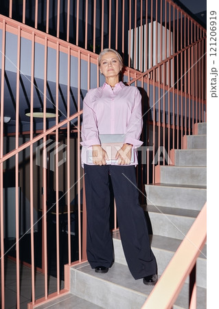 Vertical full length shot of mature businesswoman wearing pink shirt outfit confidently posing for camera standing on staircase in office building, camera flash, copy space Vertical full length shot of mature businesswoman wearing pink shirt outfit confidently posing for camera standing on staircase in office building, camera flash, copy space 121206919