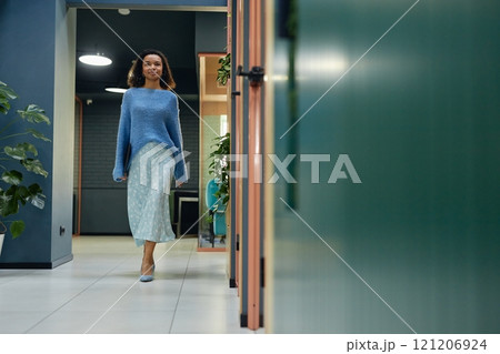 Full length shot of young curly woman in elegant blue clothes walking confidently along office rooms in corridor of modern coworking space, copy space Full length shot of young curly woman in elegant blue clothes walking confidently along office rooms in corridor of modern coworking space, copy space 121206924