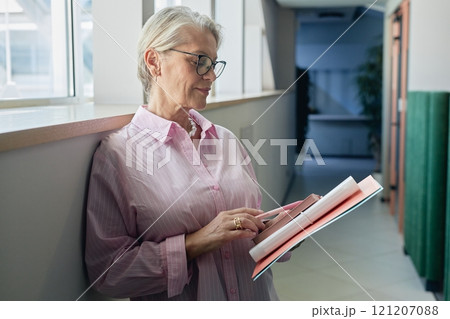 Medium shot of successful senior businesswoman wearing glasses reading note in notebook while standing in hallway of office building, copy space 121207088