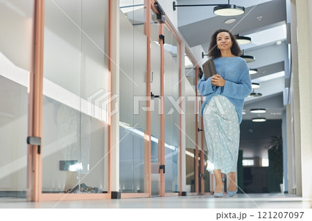 Full length shot of young multiethnic woman in elegant blue outfit holding laptop in hands walking along office rooms in hallway of modern coworking space, copy space 121207097