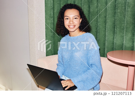 Portrait of curly multiethnic female manager wearing blue sweater outfit smiling at camera working on laptop while sitting in lounge area of modern coworking space, camera flash, copy space Portrait of curly multiethnic female manager wearing blue sweater outfit smiling at camera working on laptop while sitting in lounge area of modern coworking space, camera flash, copy space 121207113