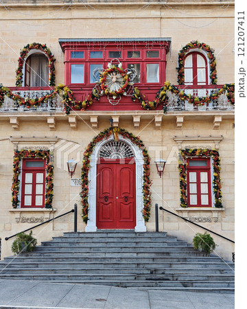 Decorated historic Maltese building with bold red door, adorned with colorful Christmas garlands and festive details Decorated historic Maltese building with bold red door, adorned with colorful Christmas garlands and festive details 121207411