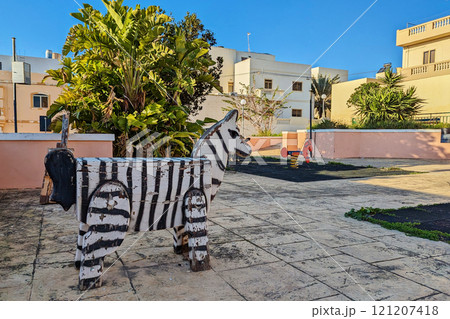 Rustic wooden zebra structure sits in weathered playground, surrounded by lush green plants in residentials area Rustic wooden zebra structure sits in weathered playground, surrounded by lush green plants in residentials area 121207418