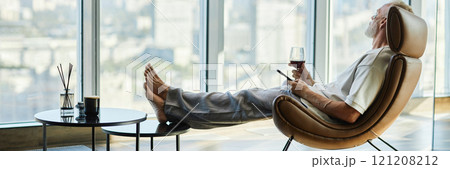 Website header shot of successful mature man leaned back in comfy armchair relaxing with glass of red wine at home 121208212