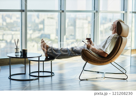 Side view of laid back successful mature man relaxing in comfy armchair with feet on footstool holding glass of red wine, while spending time in modern apartment with panoramic windows 121208230
