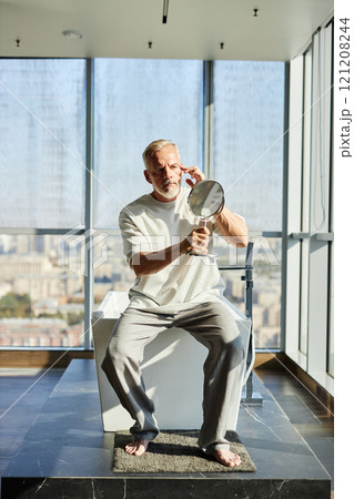 Full length shot of mature man with well groomed beard looking in hand mirror taking care of skin while sitting on bathtub in modern bathroom with panoramic windows, copy space Full length shot of mature man with well groomed beard looking in hand mirror taking care of skin while sitting on bathtub in modern bathroom with panoramic windows, copy space 121208244