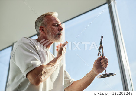 Side view of mature man with well groomed grey beard looking at reflection in mirror concerned about condition of facial hair during self care routine in bathroom, copy space Side view of mature man with well groomed grey beard looking at reflection in mirror concerned about condition of facial hair during self care routine in bathroom, copy space 121208245
