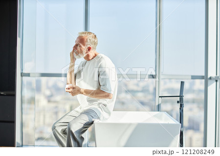 Side view of senior man with toned body applying nourishing cream on face taking care of skin while sitting on bathtub in bathroom against panoramic windows, copy space Side view of senior man with toned body applying nourishing cream on face taking care of skin while sitting on bathtub in bathroom against panoramic windows, copy space 121208249
