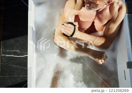 High angle shot of naked mature man taking hot foamy bath gently massaging skin with sponge during self care routine at home, copy space High angle shot of naked mature man taking hot foamy bath gently massaging skin with sponge during self care routine at home, copy space 121208269