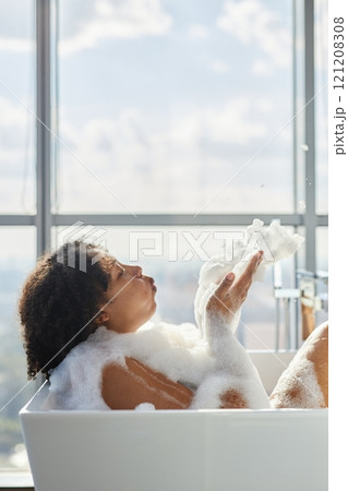 Vertical shot of happy African American woman blowing foam off palms while taking hot foamy bath in bathroom with picturesque view of city from huge windows, copy space 121208308