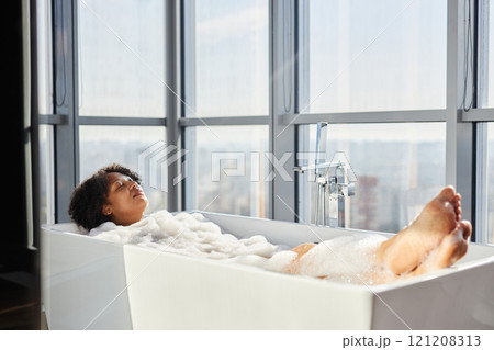Relaxed African American woman lying in bathtub enjoying hot foamy aromatic bath in room with panoramic windows, copy space 121208313