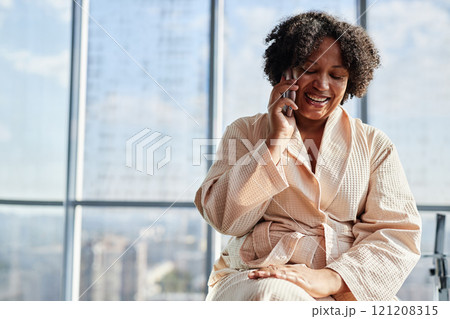 Medium shot of cheerful African American woman in comfy bathrobe talking on mobile phone during morning hygiene routine in bathroom with panoramic windows, slow life concept, copy space 121208315