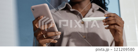 Website header shot of African American woman holding express pregnancy test while text messaging on mobile phone in bathroom 121209580