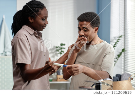 Medium shot of happy Black man holding pregnancy test looking in amazement at positive result surprised by wife at home 121209619