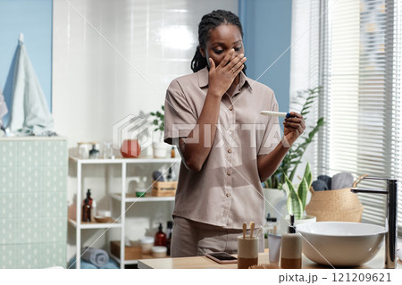 Surprised young Black woman covering face with hand in excitement looking at pregnancy test with positive result in bathroom, copy space 121209621