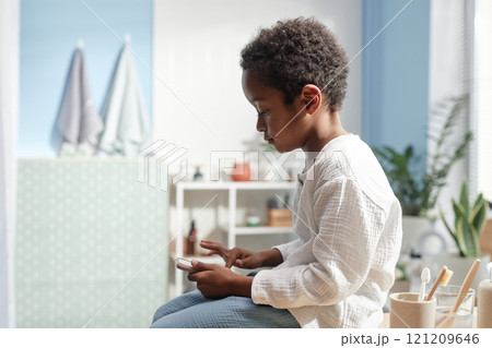 Side view of little African American boy tapping smartphone screen playing online game while sitting on tabletop in bathroom, home life scene, copy space Side view of little African American boy tapping smartphone screen playing online game while sitting on tabletop in bathroom, home life scene, copy space 121209646
