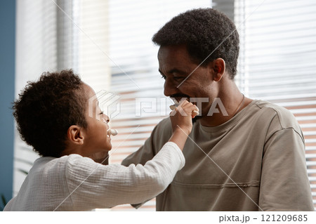 Happy Black man teaching little son dental hygiene while playful Black boy brushing fathers teeth in bathroom on Sunday morning 121209685