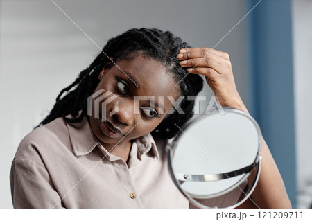 Young African American woman with natural hair looking thoroughly at reflection in mirror while examining head skin during home beauty treatment in bathroom 121209711