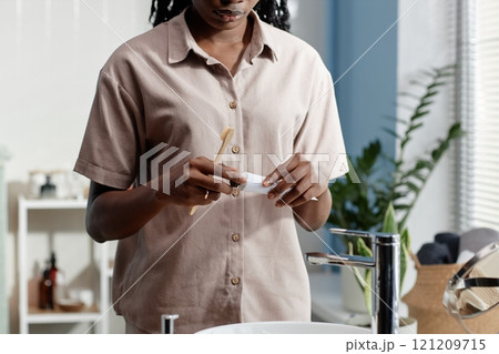 Cropped shot of young African American woman holding bamboo toothbrush and toothpaste tube taking care of dental health while standing at bathroom sink, copy space 121209715