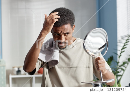 Medium shot of troubled Black man staring at reflection in mirror touching curly hair during grooming routine in bathroom, copy space 121209724