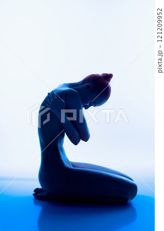 Nude figure in blue light seated with arms wrapped around knees Nude figure in blue light seated with arms wrapped around knees 121209952