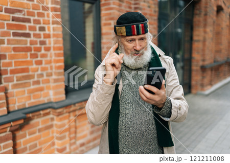 Portrait of surprised homeless elderly man having mobile phone conversation and raising index finger while standing in urban street in front of brick wall. Concept of raw reality of poverty, despair. Portrait of surprised homeless elderly man having mobile phone conversation and raising index finger while standing in urban street in front of brick wall. Concept of raw reality of poverty, despair. 121211008