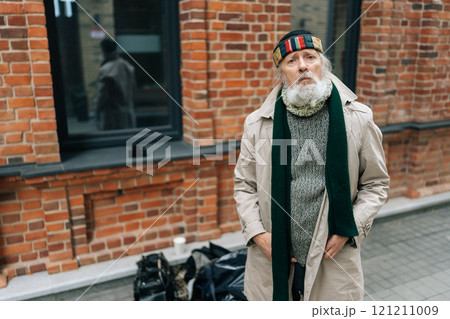 Portrait of tired homeless senior man in jacket and hat standing with hands in pockets next to red brick wall, in search of shelter or assistance, looking at camera with sadness expression. Portrait of tired homeless senior man in jacket and hat standing with hands in pockets next to red brick wall, in search of shelter or assistance, looking at camera with sadness expression. 121211009