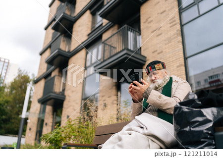 Low-angle portrait of aged homeless man sitting on city bench, using smartphone, trying to connect with family or access essential services. Concept of poverty and vulnerability, evoking empathy. Low-angle portrait of aged homeless man sitting on city bench, using smartphone, trying to connect with family or access essential services. Concept of poverty and vulnerability, evoking empathy. 121211014