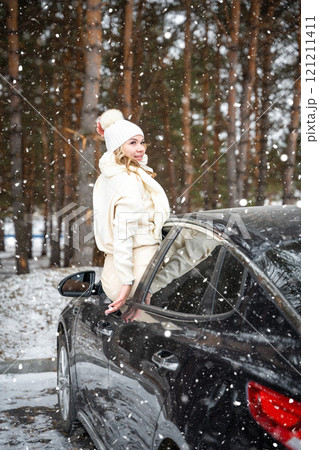 Young woman in woolen white hat have a fun in car. Winter travel, snow-covered trees in the forest. Snowy weather.  121211411