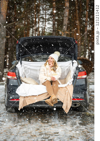Young woman in woolen hat sits in the trunk of the car. Girl have a fun in the car. Winter travel in the forest. Snowy weather.  121211412