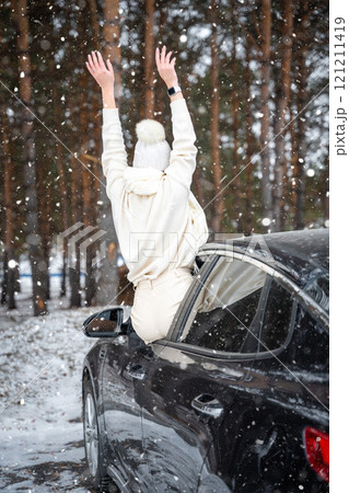 Young woman in woolen white hat have a fun in the car. Winter travel in the forest. Snowy weather.  121211419