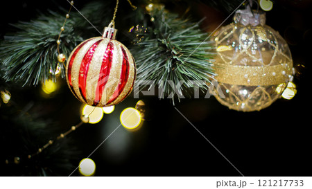 A festive close-up of two Christmas ornaments. one red and gold striped and the other elegant gold hanging from a tree with twinkling lights in the background, creating a warm holiday atmosphere. 121217733