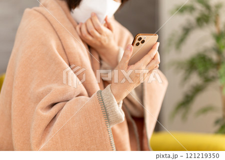 マスクをして辛そうに部屋でスマホを見るミドル〜シニア女性 121219350