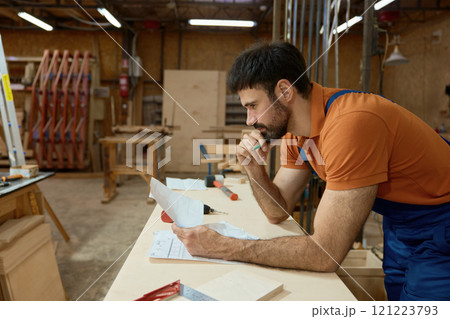 Pensive man carpenter reading blueprints and thinking during woodworking process Pensive man carpenter reading blueprints and thinking during woodworking process 121223793